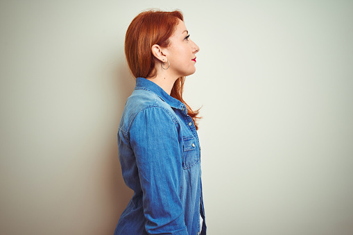 Young beautiful redhead woman wearing denim shirt standing over white isolated background looking to side, relax profile pose with natural face with confident smile.