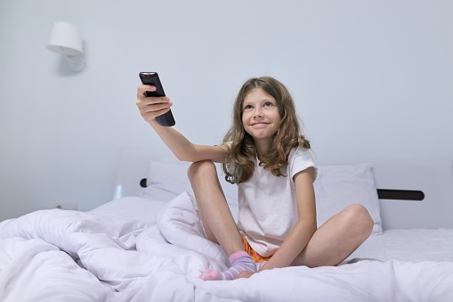 Pretty child girl blonde in morning sitting in white bed with tv remote control.