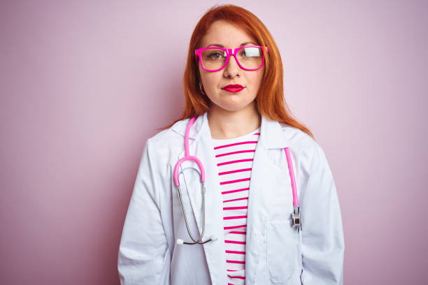 istock Young redhead doctor woman using stethoscope standing over isolated pink background with serious expression on face. Simple and natural looking at the camera. 1213948006