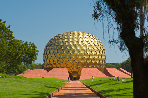 Photo of the path leading to the entrance of Matrimandir in Auroville, India. Green grass and trees surrounding the path. Blue sky behind the building. The Matrimandir is a location for meditation.