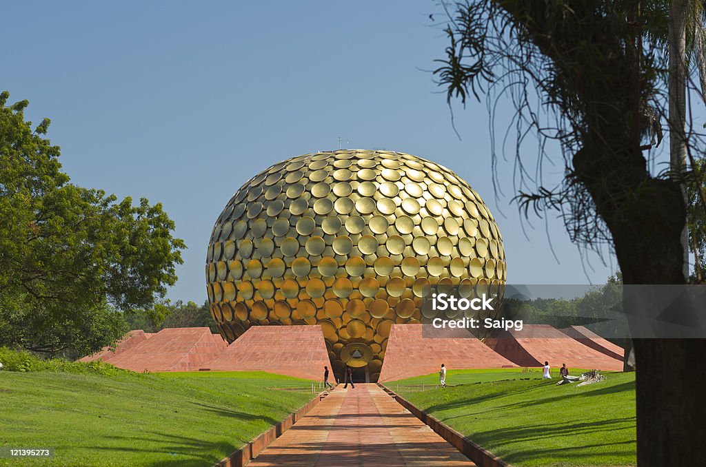 The path leading to entrance of Matrimandir in Auroville, India Photo of the path leading to the entrance of Matrimandir in Auroville, India. Green grass and trees surrounding the path. Blue sky behind the building. The Matrimandir is a location for meditation. Pondicherry Stock Photo