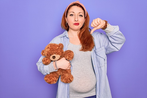 Young redhead pregnant woman expecting baby holding teddy bear over purple background with angry face, negative sign showing dislike with thumbs down, rejection concept