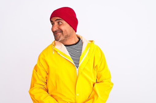 Middle age man wearing rain coat and woolen hat standing over isolated white background smiling looking to the side and staring away thinking.