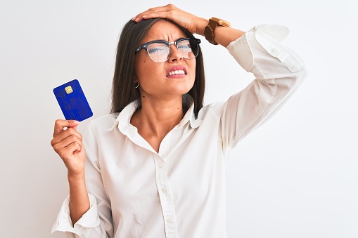 Young businesswoman wearing glasses holding credit card over isolated white background stressed with hand on head, shocked with shame and surprise face, angry and frustrated. Fear and upset for mistake.