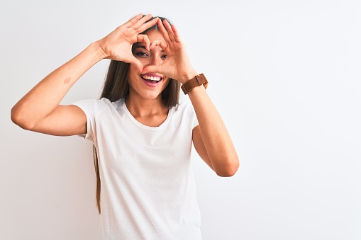 Young beautiful woman wearing casual t-shirt standing over isolated white background Doing heart shape with hand and fingers smiling looking through sign