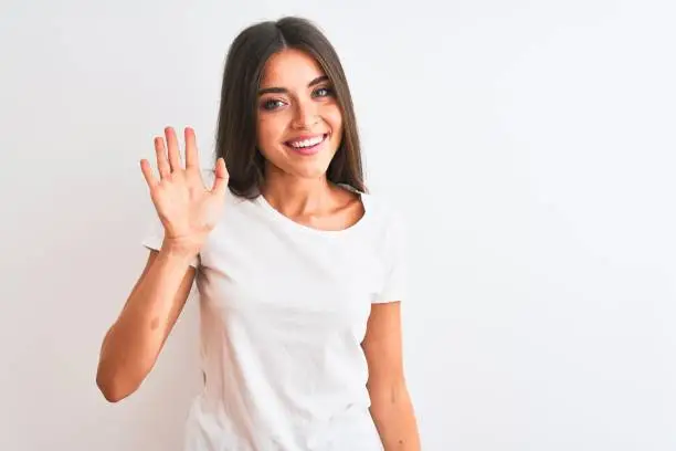 Photo of Young beautiful woman wearing casual t-shirt standing over isolated white background Waiving saying hello happy and smiling, friendly welcome gesture