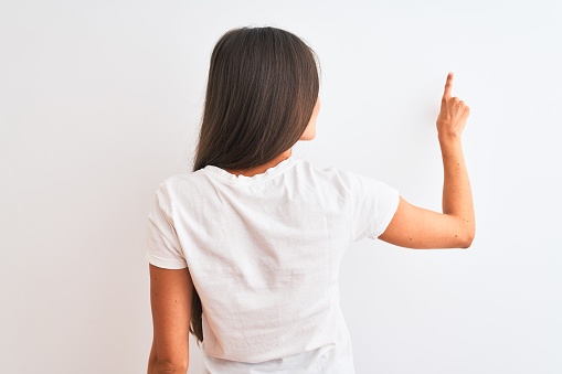 Young beautiful woman wearing casual t-shirt standing over isolated white background Posing backwards pointing ahead with finger hand