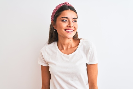 Young beautiful woman wearing casual t-shirt and diadem over isolated white background looking away to side with smile on face, natural expression. Laughing confident.