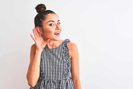 Beautiful woman with bun wearing casual dresss standing over isolated white background smiling with hand over ear listening an hearing to rumor or gossip. Deafness concept.