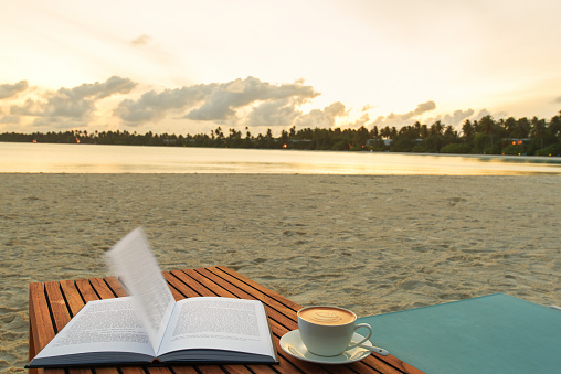 Open book and coffee cup on beach table beside tropical sea at sunrise time