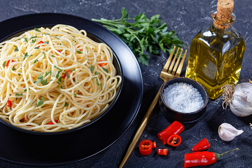Pasta Aglio, Olio e Peperoncino, italian spaghetti with garlic, chili and olive oil in a black bowl on a concrete table with ingredients on a table, horizontal view from above, close-up