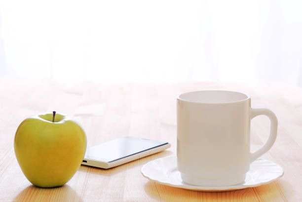 white coffee cup, green apple and smartphone lit by morning sunlight on a wooden table, copy space - wakening imagens e fotografias de stock