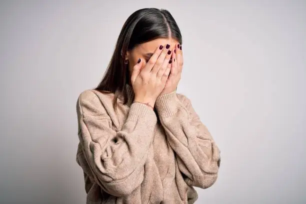 Photo of Young beautiful brunette woman wearing casual sweater standing over white background with sad expression covering face with hands while crying. Depression concept.