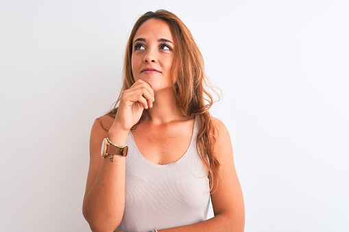 Young redhead woman wearing casual t-shirt stading over white isolated background with hand on chin thinking about question, pensive expression. Smiling with thoughtful face. Doubt concept.