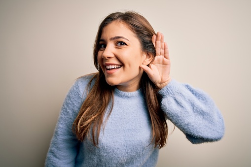 Beautiful young woman wearing casual winter sweater standing over isolated background smiling with hand over ear listening an hearing to rumor or gossip. Deafness concept.