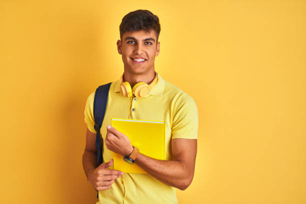 istock Indian student man wearing backpack headphones notebook over isolated yellow background with a happy face standing and smiling with a confident smile showing teeth 1214024597