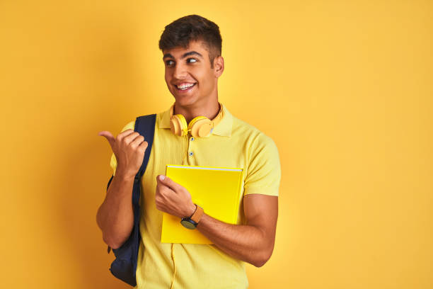 istock Indian student man wearing backpack headphones notebook over isolated yellow background pointing and showing with thumb up to the side with happy face smiling 1214024620