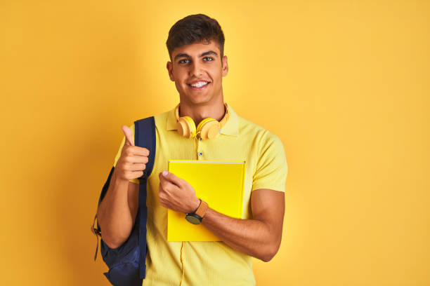istock Indian student man wearing backpack headphones notebook over isolated yellow background happy with big smile doing ok sign, thumb up with fingers, excellent sign 1214024640