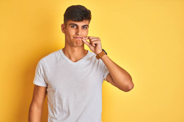 고립 된 노란색 배경 입과 입술 위에 서있는 흰색 티셔츠를 입은 젊은 인도 남자가 손가락으로 지퍼로 닫습니다. ��비밀과 침묵, 금기 이야기 - taboo 뉴스 사진 이미지