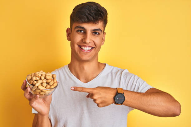 jeune homme indien retenant le bol avec des arachides au-dessus du fond jaune isolé très heureux pointant avec la main et le doigt - indian nut photos et images de collection