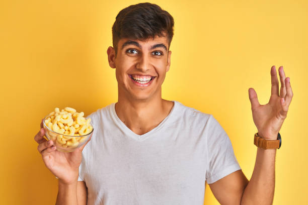 istock Young indian man holding bowl with extruded corn standing over isolated yellow background very happy and excited, winner expression celebrating victory screaming with big smile and raised hands 1214026962