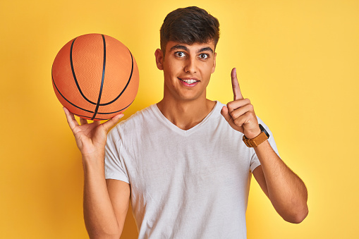 Young indian sportsman holding basketball ball standing over isolated yellow background surprised with an idea or question pointing finger with happy face, number one