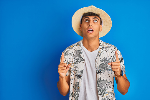 Indian man on vacation wearing hawiaian shirt summer hat over isolated blue background amazed and surprised looking up and pointing with fingers and raised arms.