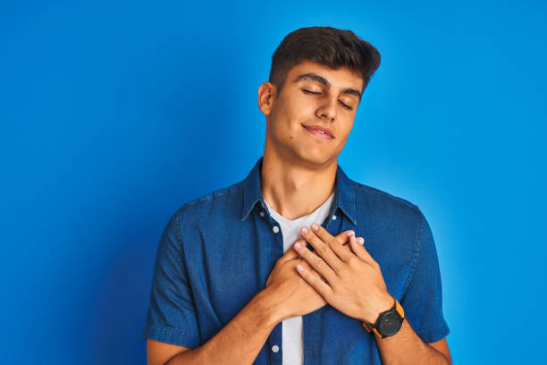 istock Young indian man wearing shirt standing over isolated blue background smiling with hands on chest with closed eyes and grateful gesture on face. Health concept. 1214029556
