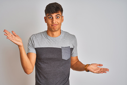 Young indian man wearing casual t-shirt standing over isolated white background clueless and confused expression with arms and hands raised. Doubt concept.