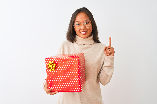 Young chinese woman wearing glasses holding birthday gift over isolated white background surprised with an idea or question pointing finger with happy face, number one