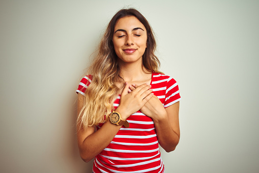 Young beautiful woman wearing red stripes t-shirt over white isolated background smiling with hands on chest with closed eyes and grateful gesture on face. Health concept.