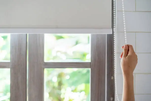 Cropped view of woman pull rope, opened modern white blinds roll on windows in light and tiled bathroom