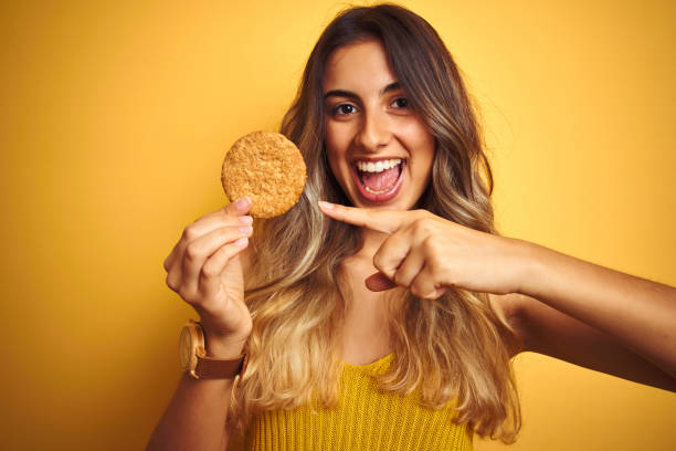 jovem mulher bonita comendo biscoito sobre cinza fundo isolado muito feliz apontando com a mão e o dedo - cookie women eating beautiful - fotografias e filmes do acervo