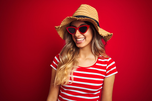 Young beautiful woman wearing sunglasses and summer hat over red isolated background looking away to side with smile on face, natural expression. Laughing confident.