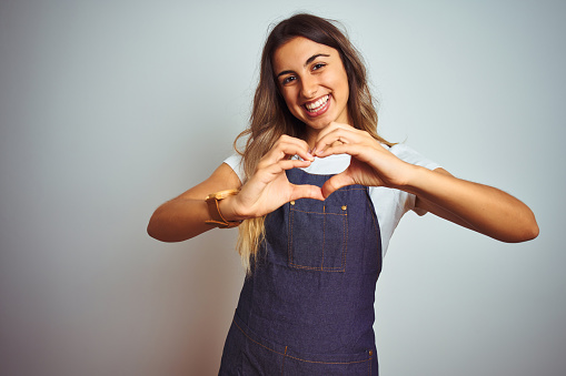 Young beautiful woman wearing apron over grey isolated background smiling in love showing heart symbol and shape with hands. Romantic concept.