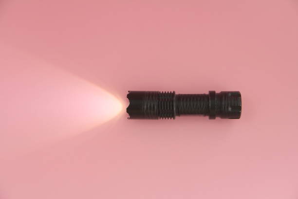 la lampada manuale elettrica accesa in nero giace su uno sfondo rosa. luce da una lanterna sulla superficie - tactical flashlight foto e immagini stock