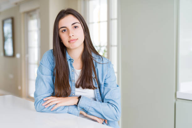 istock Beautiful young woman sitting on white table at home with serious expression on face. Simple and natural looking at the camera. 1214054955