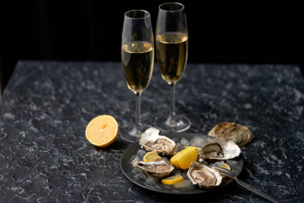 open oesters op een zwarte schotel met plakken van verpletterd ijs, citroenplakken en twee glazen die met witte wijn op een zwarte steen geweven achtergrond worden gevuld. bovenste weergave onder een hoek. plaats voor tekst - andrej stockfoto's en -beelden