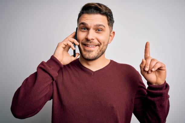 istock Young man with blue eyes speaking on the phone having a conversation on smartphone surprised with an idea or question pointing finger with happy face, number one 1214057190