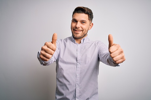 Young business man with blue eyes standing over isolated background approving doing positive gesture with hand, thumbs up smiling and happy for success. Winner gesture.