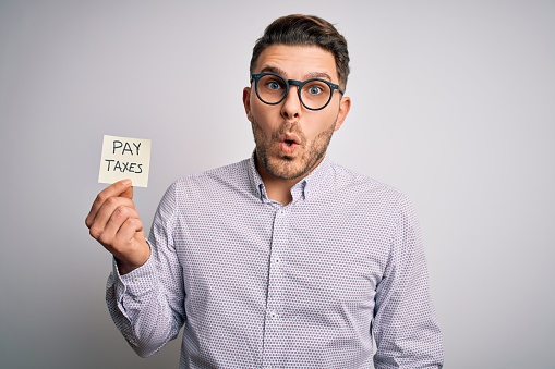 Young business man with blue eyes holding pay taxes word on paper note scared in shock with a surprise face, afraid and excited with fear expression