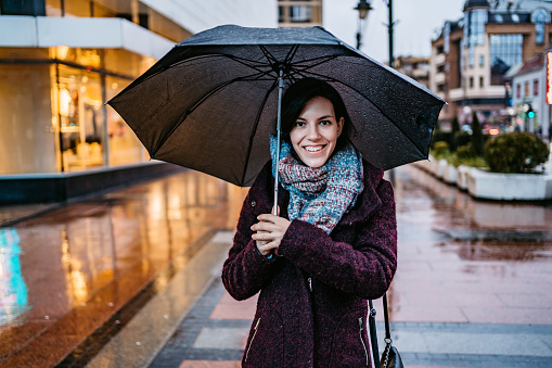 Young Caucasian smiling woman walking downtown in rain with umbrella. Autumn time.