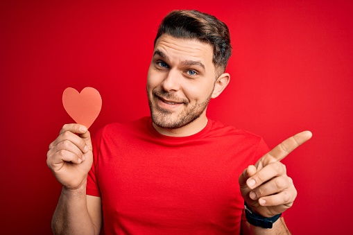 Young romantic man with blue eyes holding red heart paper shaped over red background very happy pointing with hand and finger to the side