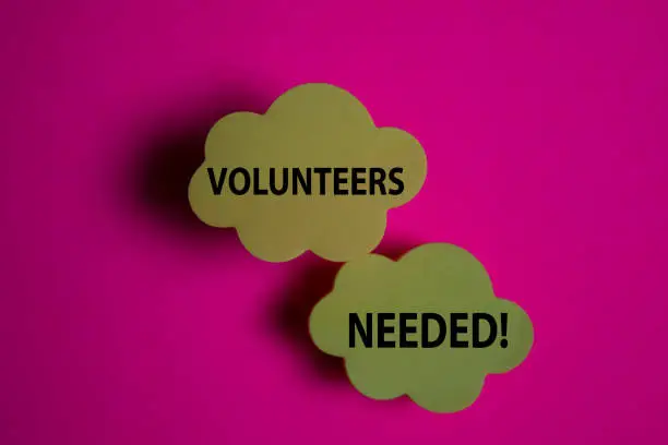 Photo of Volunteers Needed! write on a sticky note isolated on Office Desk.