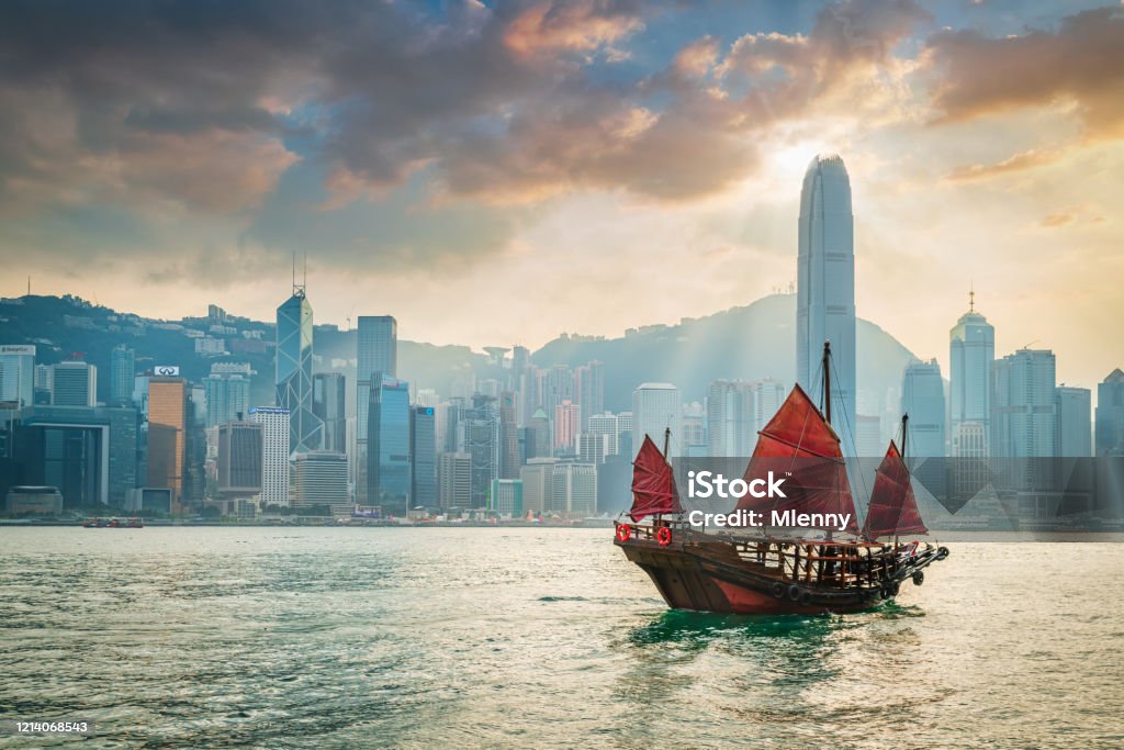 Red Sail Junkboat Cruising Along Hong Kong Cityscape at Sunset Traditional chinese junkboat with typical red sails sailing across victoria harbour under colurful amazing sunset twilight and cloudscape. Hong Kong Skyline in the Background, Hong Kong, Southeast Asia. Hong Kong Stock Photo