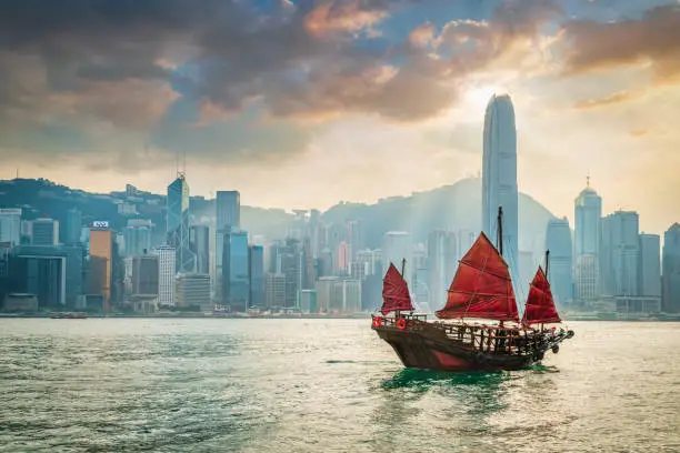 Traditional chinese junkboat with typical red sails sailing across victoria harbour under colurful amazing sunset twilight and cloudscape. Hong Kong Skyline in the Background, Hong Kong, Southeast Asia.