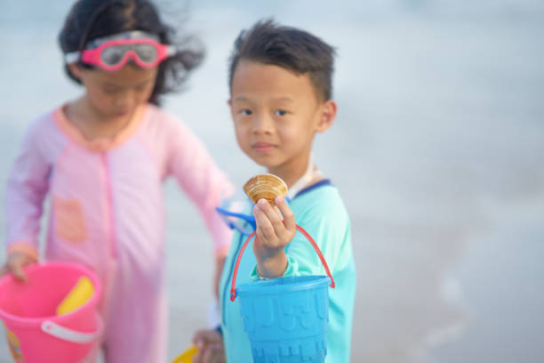 Bare legs in crocs stand at the edge of a rocky shore, as playful waves lap at the feet. A bright yellow bucket swings gently in hand, ready to collect treasures from the sea. This close-up frame captures the pure joy and sensory richness of childhood by the water  the sound of waves, the splash of foam, and the promise of discovery.