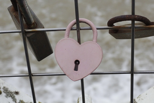 Love heart shape, aged Pink padlock, attached to silver metal wire fence, in front of frothy water foam with Old gold lock surrounding it