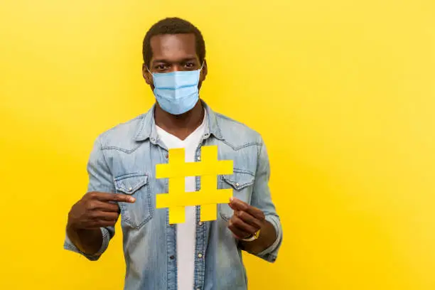 Photo of Viral hashtag and successful blogging. Portrait of happy man with attractive toothy smile with medical mask pointing at large yellow hash symbol.