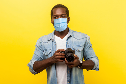Portrait of positive photographer, man with medical mask holding professional digital dslr camera and looking with toothy smile, enjoying his job.